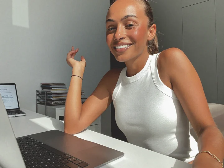 Jamie R. smiling at her desk beside an open laptop in bright morning light.