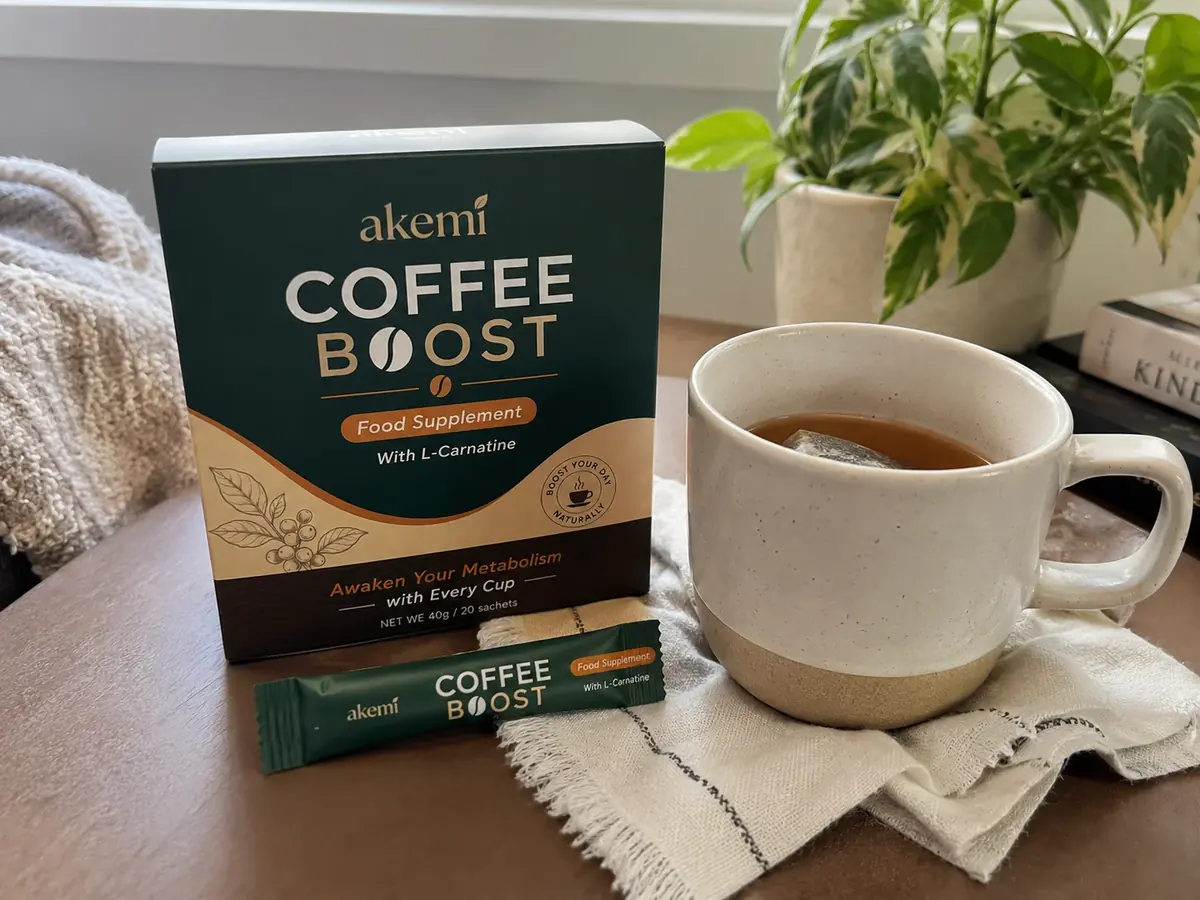 The Akemi Coffee Boost carton and a single sachet on a wooden table beside a stoneware mug of black coffee